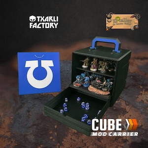 Op de afbeelding: Een donkergroene Cube Mod Carrier met een blauwe handgreep en een uitschuifbare lade. De carrier is gevuld met miniatuurbeeldjes en blauwe dobbelstenen. Links staat een blauw bord met een wit symbool. De tekst "TXARLI FACTORY" staat bovenaan.