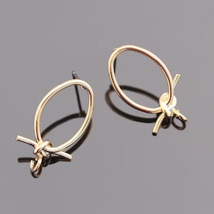 May include: A pair of gold-colored earrings. Each earring features an oval loop design with a knot detail at the bottom. The earrings have a post backing for pierced ears. The jewelry is displayed on a dark surface.