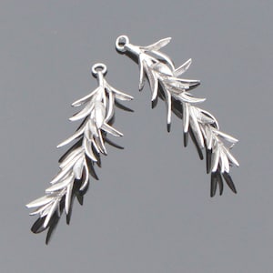 Puede incluir: Dos colgantes plateados con forma de hoja. Cada colgante tiene un lazo en la parte superior para colgar y un diseño detallado y texturizado de hojas y ramas. Los colgantes se muestran sobre un fondo gris.