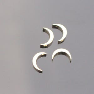 May include: Four small, gold-colored crescent moon charms are arranged on a reflective, dark gray surface. Each charm has a curved shape with a small hole at each end, suggesting they are designed for jewelry making or crafting.