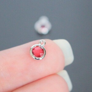 May include: A close-up of a silver charm with a red rhinestone. The charm is round and has a loop at the top for attaching to a bracelet or necklace.