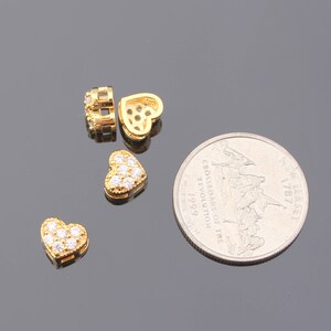May include: Four gold-tone heart-shaped charms with clear crystal accents. The charms are on a gray surface next to a US quarter.
