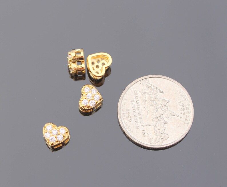 May include: Four gold-tone heart-shaped charms with clear crystal accents. The charms are on a gray surface next to a US quarter.