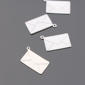 May include: Several silver-colored envelope charms with a loop at the top for jewelry making. The charms are rectangular with a detailed envelope design. The background is a solid gray color.