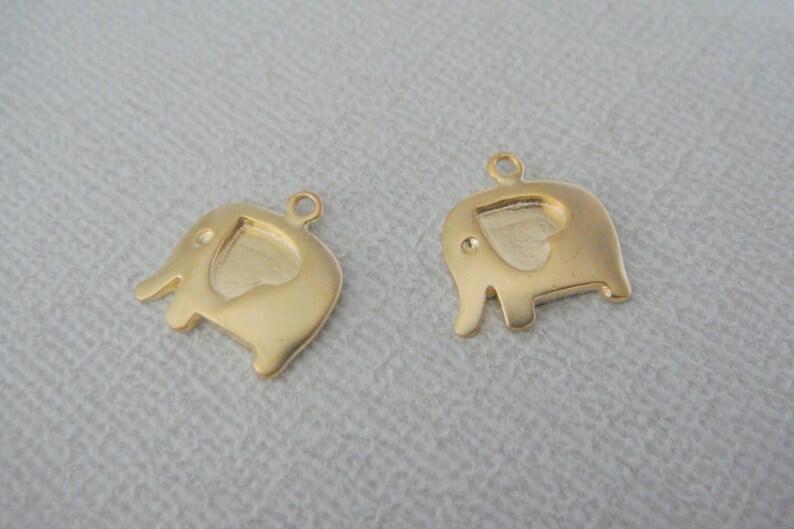 May include: Two gold-colored elephant charms with a heart-shaped cutout in the center. The charms have a loop at the top for attaching to a necklace or bracelet.