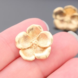 May include: Close-up of a gold-colored flower-shaped jewelry component. The component has five petals with detailed veining and a textured center. Another similar component is slightly out of focus in the background.