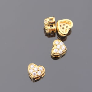 May include: Four gold heart-shaped charms with clear cubic zirconia stones. The charms are on a gray surface.