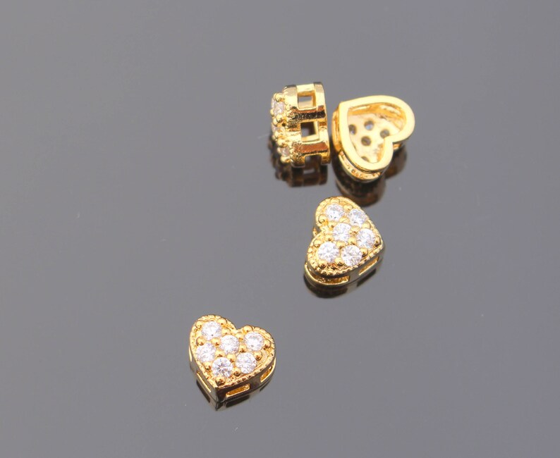 May include: Four gold heart-shaped charms with clear cubic zirconia stones. The charms are on a gray surface.
