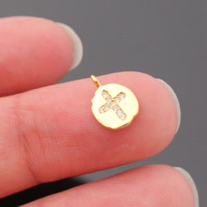 May include: Gold charm with a cross design, featuring small clear rhinestones. The charm is on a finger.