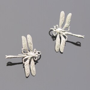 May include: Two silver dragonfly pendants. Each pendant has detailed wings and a slender body. The dragonflies are on a dark grey surface, casting shadows. The pendants are likely for jewellery making or as decorative elements.