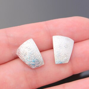 May include: Two silver-colored, curved metal components with a textured, swirling pattern. The pieces are small and appear to be jewelry components, possibly for earrings or other accessories. The background is a neutral gray.