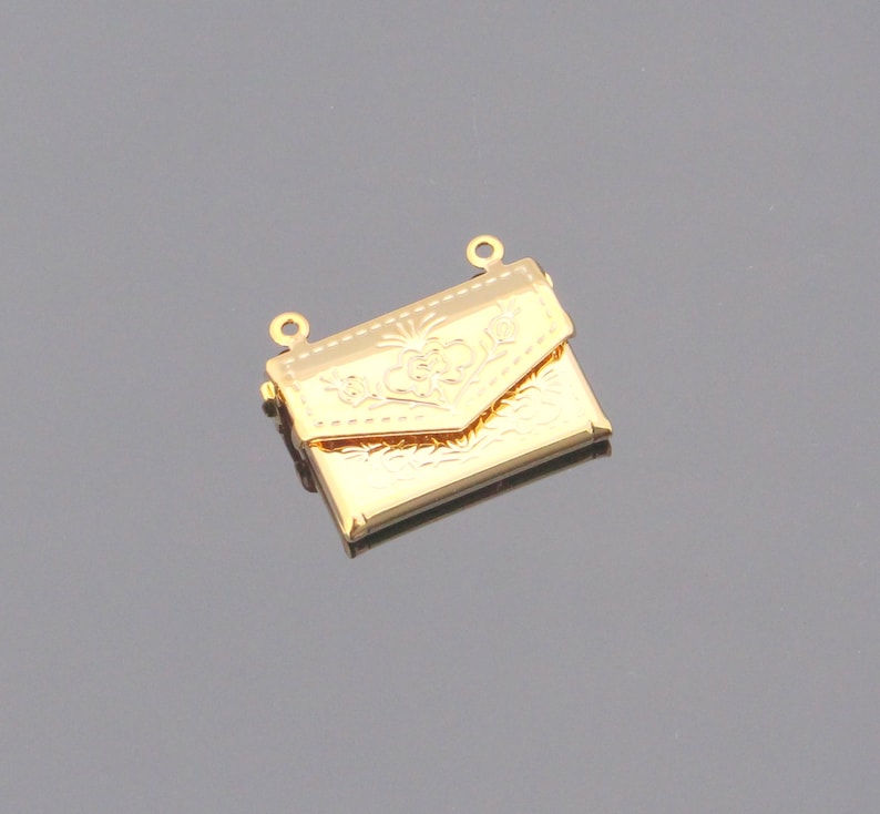 May include: Gold-toned metal charm in the shape of a small, ornate envelope with a floral design. The charm has two loops at the top for attaching to a necklace or bracelet.