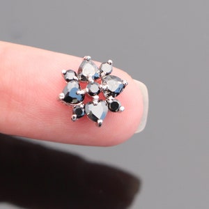 以下が含まれることがあります： 黒いキュービックジルコニアの石がセットされた、花の形をしたシルバーの鼻のリングです。