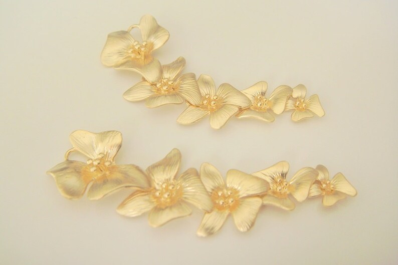 May include: Two gold-toned flower-shaped hair clips. Each clip features a series of five connected flowers with a textured surface.