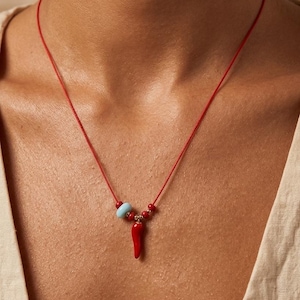 Può includere: Collana con cordino rosso con un piccolo ciondolo a forma di peperoncino rosso. La collana include una perlina azzurra e diverse piccole perline rosse. La collana è indossata intorno al collo.