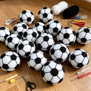 May include: A collection of fifteen handmade crocheted soccer balls featuring a classic white and black pentagon pattern. The set includes various crochet hooks, scissors, and skeins of white and black yarn arranged around the spheres.