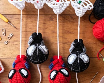 May include: Four crocheted ornaments featuring two red and blue superhero figures and two black and white spherical figures, each suspended by a white yarn string from a white crocheted net structure with a small colored bead at the center.