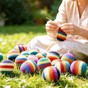 May include: A collection of colorful, crocheted balls with rainbow stripes, resting on green grass. The balls are in various colors, including red, orange, yellow, green, blue, and purple. A person is crocheting a similar ball in the background.