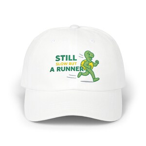 May include: White baseball cap with the text "STILL SLOW BUT A RUNNER" in green. A cartoon turtle in green and yellow is running to the right. The cap has a curved brim and a structured fit.