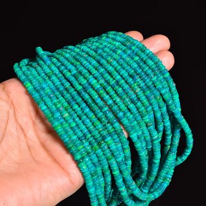 May include: Strands of small, round, teal-colored beads. The beads have a slight iridescence, with hints of green and yellow. The beads are held in a hand against a black background. The beads appear to be strung together, forming multiple strands.