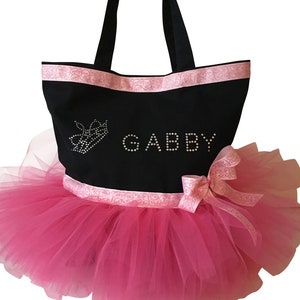 May include: Black tote bag with pink ribbon trim and the name "GABBY" spelled out in rhinestones. A pink tutu skirt is attached to the bottom of the bag, with a matching pink bow. A rhinestone crown is also on the bag.