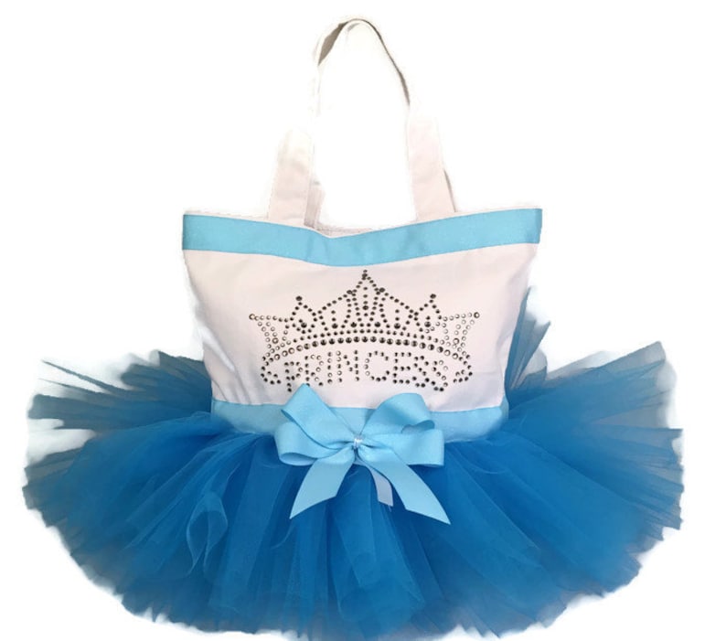 Peut inclure: Un sac fourre-tout blanc avec une bordure et des poign&eacute;es bleu clair, orn&eacute; d'une couronne en strass et du mot "PRINCESS". Il est associ&eacute; &agrave; une jupe tutu en tulle bleu et un n&oelig;ud assorti.