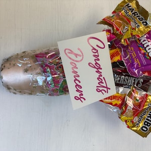May include: A ballet slipper wrapped in clear cellophane, adorned with gold stars, and a collection of candy. A white card with the words "Congrats Dancers" in pink script is included.