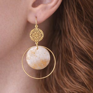 Könnte beinhalten: Goldfarbene Ohrringe mit kreisförmigem Design. Die Ohrringe verfügen über eine kleine, aufwendig gemusterte Scheibe über einem größeren, weiß und goldfarben gesprenkelten Kreis, der in einem goldenen Reifen aufgehängt ist. Die Ohrringe werden vor einem verschwommenen Hintergrund gezeigt.