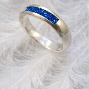 Può includere: Un anello d'argento con un intarsio rettangolare di materiale blu scintillante. L'anello è posto su uno sfondo bianco e morbido di piume, creando un contrasto tra il metallo freddo e la delicata consistenza.