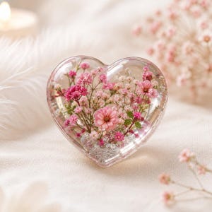 以下が含まれることがあります： 保存されたピンクの花と小さな緑の茎が入った、ハート型の透明な樹脂製ペーパーウェイト。内部には、透明な素材の中に繊細なピンクの花と小さなつぼみの群れが閉じ込められており、花のディスプレイを作り出しています。