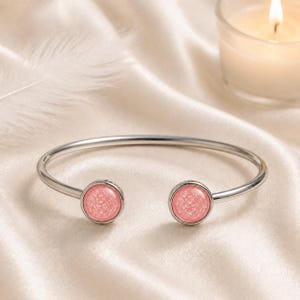 May include: A silver-toned open cuff bracelet featuring two circular end caps, each inlaid with a textured, light pink, glittery material. The bracelet has a smooth, polished metal band that curves into a circular shape with an opening at the front.