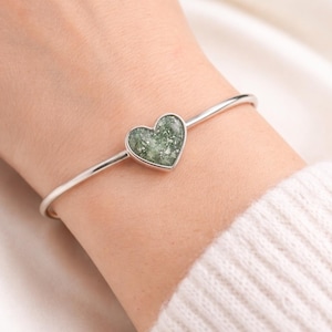 Puede incluir: Una pulsera de brazalete plateada con un dije en forma de corazón. El corazón está relleno de un material verde moteado. La pulsera se lleva en la muñeca, con un suéter blanco en el fondo.