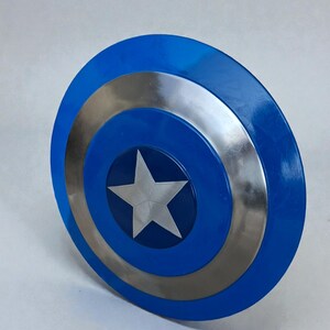May include: A blue and silver shield with a white star in the center. The shield has a circular design with a blue outer ring, a silver middle ring, and a blue center with a white star.
