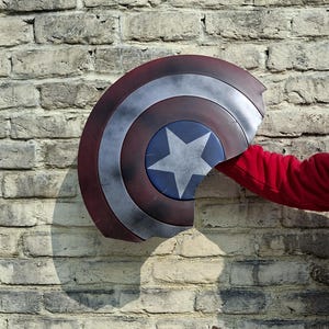 May include: A Captain America shield with a red, white, and blue color scheme. The shield has a star in the center and is held against a brick wall. The shield is approximately 24 inches in diameter.