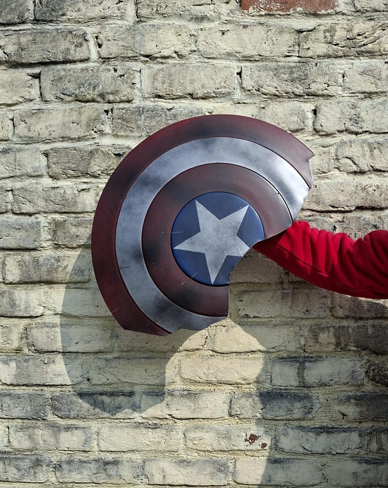 May include: A Captain America shield with a red, white, and blue color scheme. The shield has a star in the center and is held against a brick wall. The shield is approximately 24 inches in diameter.