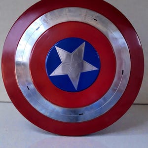 Puede incluir: Un escudo redondo con un anillo exterior rojo, una banda plateada y un centro azul. Una estrella plateada est&aacute; en el centro del c&iacute;rculo azul. El escudo es una r&eacute;plica del escudo del Capit&aacute;n Am&eacute;rica.