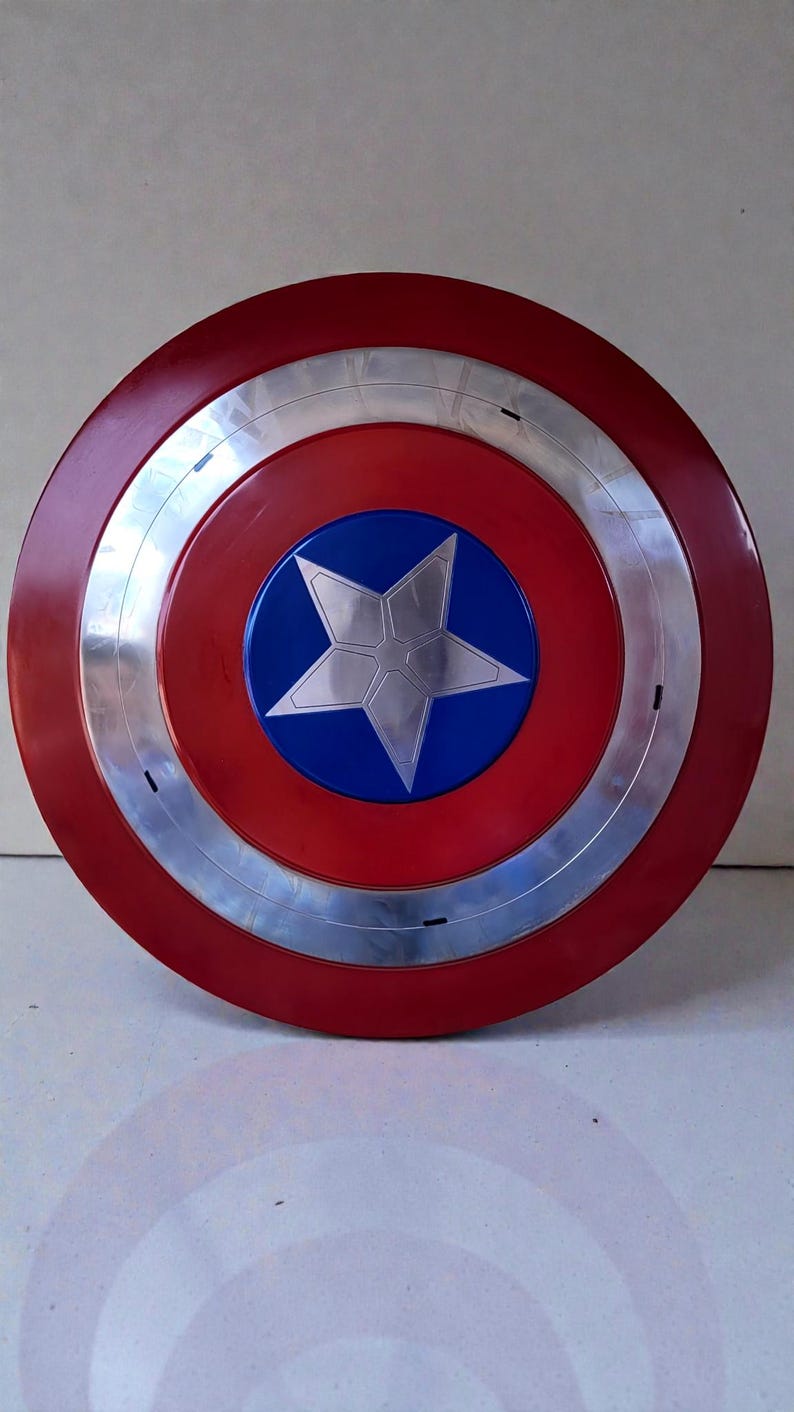 Puede incluir: Un escudo redondo con un anillo exterior rojo, una banda plateada y un centro azul. Una estrella plateada est&aacute; en el centro del c&iacute;rculo azul. El escudo es una r&eacute;plica del escudo del Capit&aacute;n Am&eacute;rica.