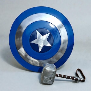 May include: A blue and silver Captain America shield with a white star in the center. Next to it is a silver hammer with a brown handle and a leather strap. Superhero themed props.