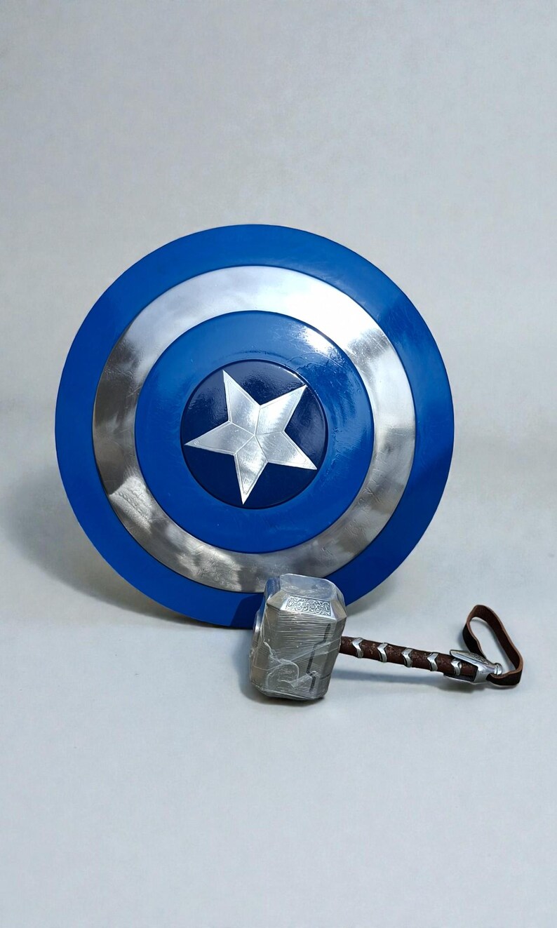 May include: A blue and silver Captain America shield with a white star in the center. Next to it is a silver hammer with a brown handle and a leather strap. Superhero themed props.