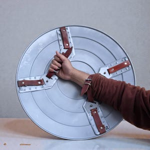 May include: A silver circular shield with concentric rings. It features four brown leather straps secured with rivets. The shield is held by a person.