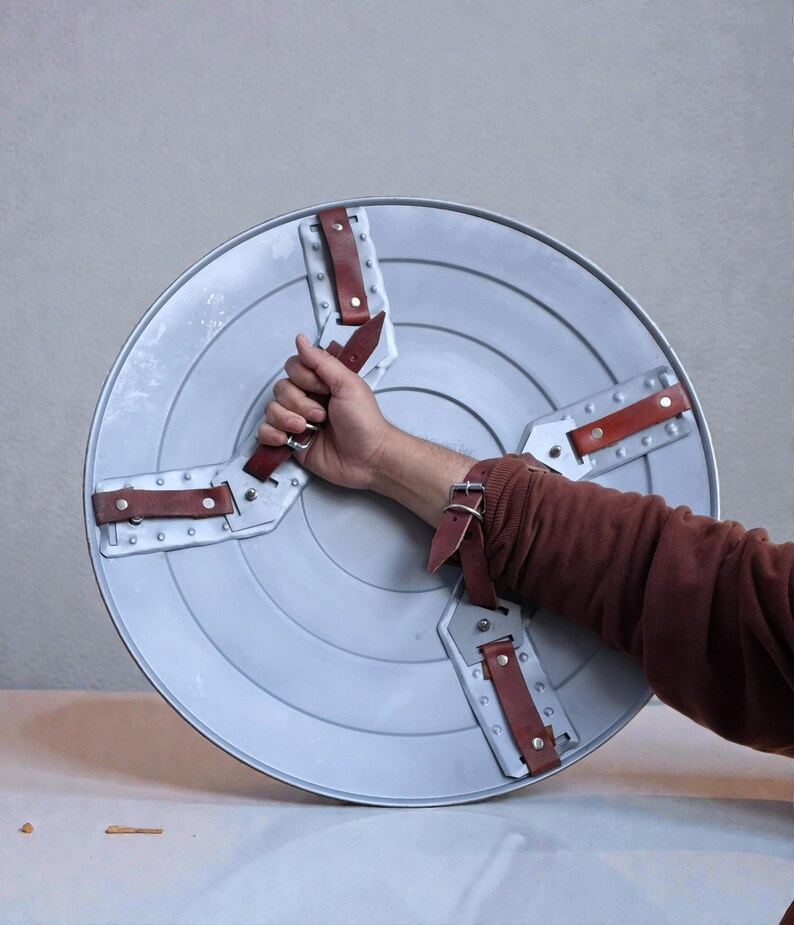 May include: A silver circular shield with concentric rings. It features four brown leather straps secured with rivets. The shield is held by a person.
