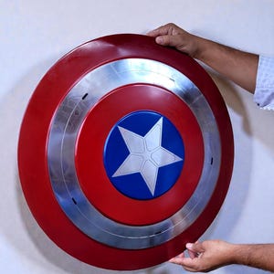 Puede incluir: Un escudo del Capit&aacute;n Am&eacute;rica rojo, plateado y azul. El escudo es redondo con un anillo plateado y un centro azul con una estrella blanca. El escudo se sostiene contra una pared blanca.