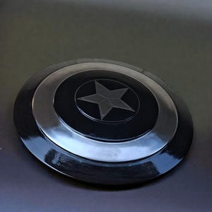 May include: A black and silver shield featuring a central star design. The shield is circular with concentric rings and a glossy finish. The star is a five-pointed design in the centre of the shield.