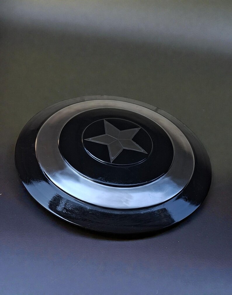 May include: A black and silver shield featuring a central star design. The shield is circular with concentric rings and a glossy finish. The star is a five-pointed design in the centre of the shield.