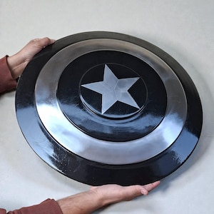 May include: A black and silver shield featuring a central white star. The shield is circular with concentric rings of black and silver. The item is held by two hands.