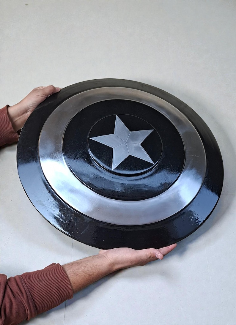 May include: A black and silver shield featuring a central white star. The shield is circular with concentric rings of black and silver. The item is held by two hands.