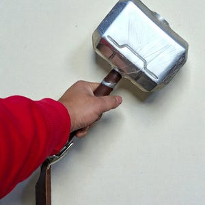 May include: A silver-colored hammer with a brown handle and a leather strap. The hammer's head has a rectangular shape with etched details. The handle is wrapped with a silver band. The hammer is held by a person wearing a red shirt.