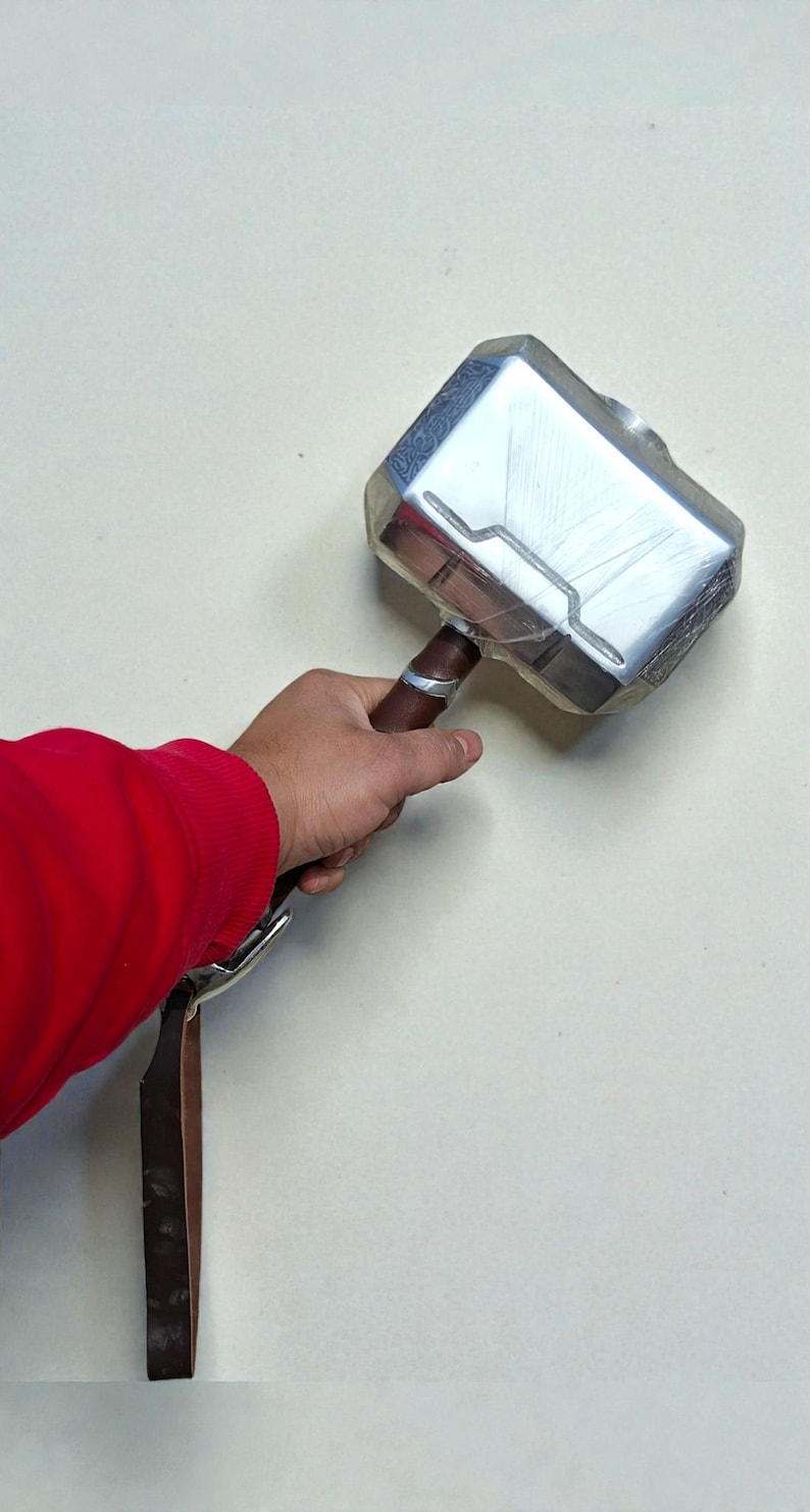 May include: A silver-colored hammer with a brown handle and a leather strap. The hammer's head has a rectangular shape with etched details. The handle is wrapped with a silver band. The hammer is held by a person wearing a red shirt.