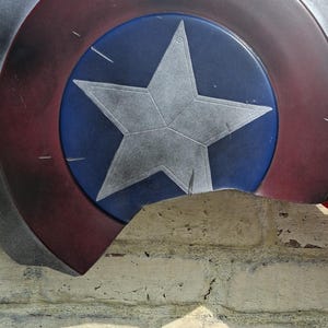 May include: A Captain America shield, featuring a blue central disc with a white star, surrounded by concentric rings of red, white, and silver. The shield is partially visible against a textured brick wall.