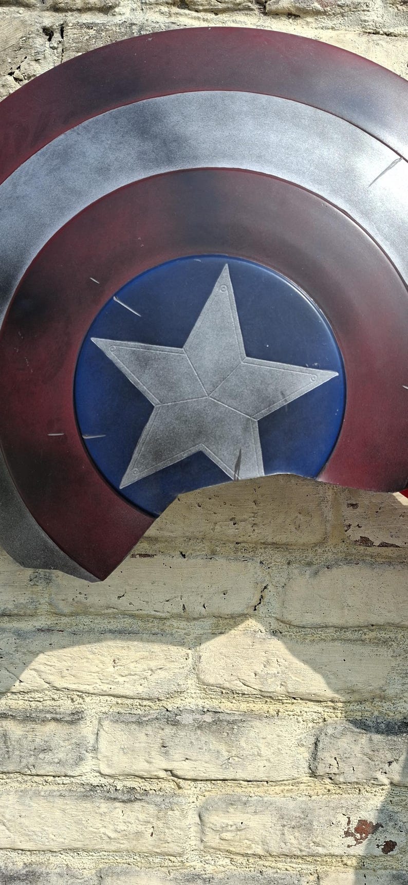 May include: A Captain America shield, featuring a blue central disc with a white star, surrounded by concentric rings of red, white, and silver. The shield is partially visible against a textured brick wall.